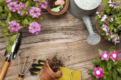 A close-up view of a wooden garden table with various gardening tools and plant pots. Included are a small trowel with a black handle, a hand rake with a wooden handle, a pair of gardening gloves with yellow cuffs, and a terracotta pot filled with soil and small plant labels. Surrounding the tools are potted flowering plants with vibrant purple and pink blossoms, some in decorative containers made of clay and metal. The table surface has a weathered appearance, showing natural wood grain and slight wear marks, and the scene is set outdoors under natural daylight, suggesting a well-maintained garden space in Kings Cross or nearby London. This setup reflects the preparation stages for planting or garden maintenance, typical of professional gardening services offered by Gardening Services Kings Cross, focusing on outdoor cultivation and landscape care.
