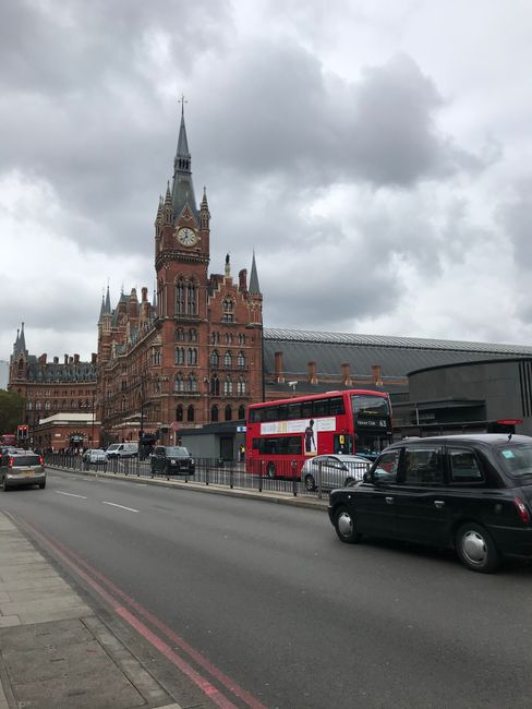 A street scene in front of a large, historic red-brick building with Gothic architectural features, including a tall clock tower with a spire and intricate detailing, set against a cloudy sky. The for