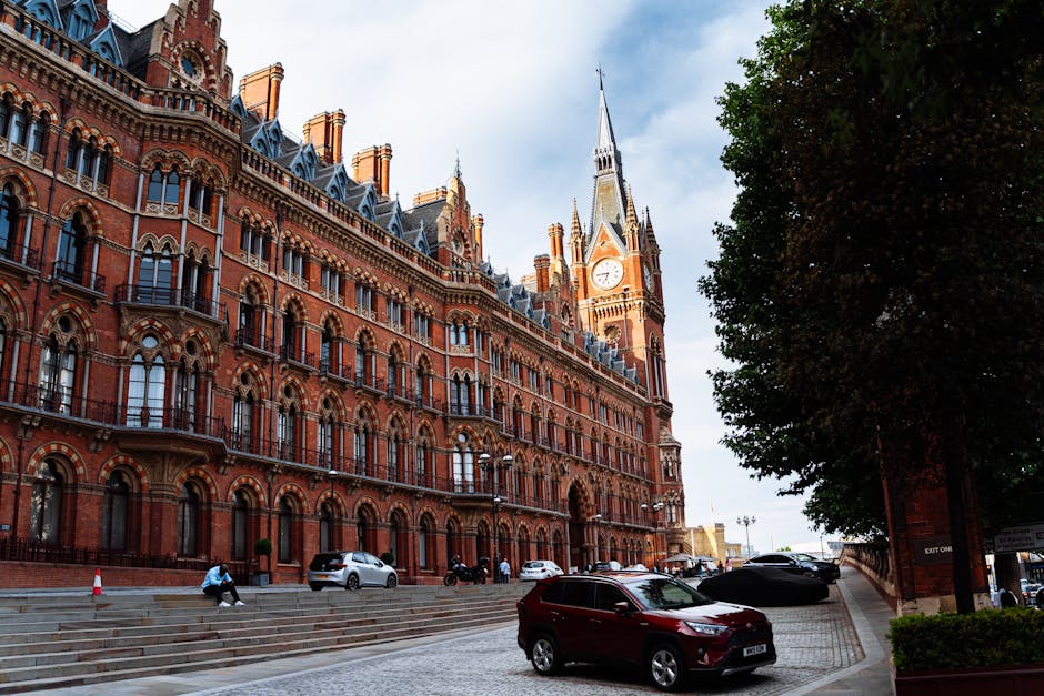 A street scene in front of a large, historic red-brick building with Gothic architectural features, including a tall clock tower with a spire and intricate detailing, set against a cloudy sky. The for