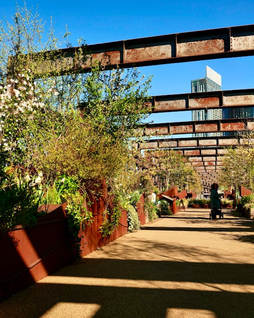 A landscaped urban garden situated beneath a series of rust-colored metal overhead structures, featuring a variety of lush green shrubs, small trees, and flowering plants in large, matching rust-color