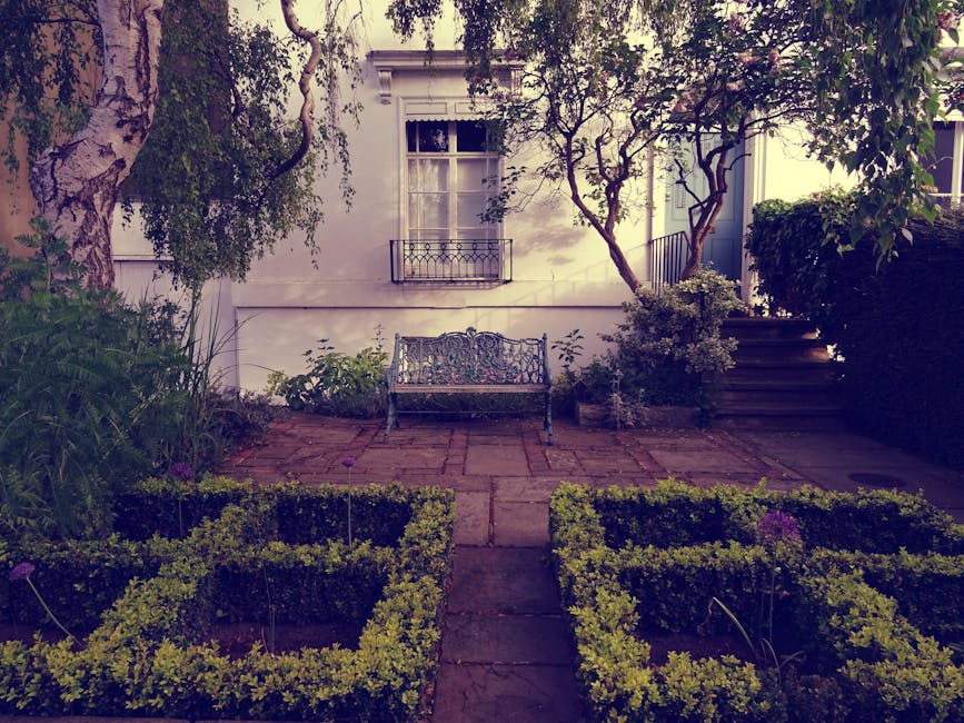 A landscaped urban garden situated beneath a series of rust-colored metal overhead structures, featuring a variety of lush green shrubs, small trees, and flowering plants in large, matching rust-color